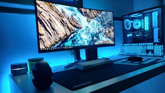 A modern computer setup featuring a large monitor displaying an ocean scene with rocks, surrounded by LED lighting in a calming blue hue. The desktop is sleek and minimalistic, with a mechanical keyboard, a gaming mouse, and headphones resting on a white table. The computer tower is visible with components illuminated by matching blue LED lights.