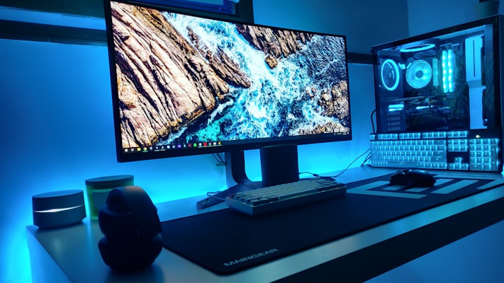 A modern computer setup featuring a large monitor displaying an ocean scene with rocks, surrounded by LED lighting in a calming blue hue. The desktop is sleek and minimalistic, with a mechanical keyboard, a gaming mouse, and headphones resting on a white table. The computer tower is visible with components illuminated by matching blue LED lights.