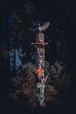 A detailed totem pole stands prominently against a backdrop of dense, dark green foliage. The pole is intricately carved and painted with vibrant colors, depicting various figures and symbols. The top figure resembles a bird with outstretched wings, while other figures include human and animal shapes. The craftsmanship reflects a rich cultural heritage.