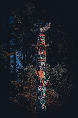 A detailed totem pole stands prominently against a backdrop of dense, dark green foliage. The pole is intricately carved and painted with vibrant colors, depicting various figures and symbols. The top figure resembles a bird with outstretched wings, while other figures include human and animal shapes. The craftsmanship reflects a rich cultural heritage.