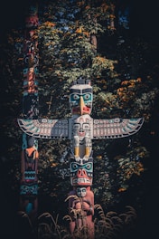 A traditional totem pole standing prominently against a backdrop of dense green foliage with accents of yellow and orange. The intricately carved wooden structure features vibrant colors and multiple figures, including a large bird with outstretched wings at the top and various human and animal faces below.