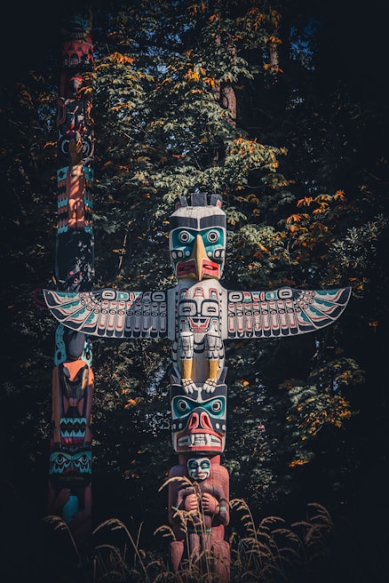 A traditional totem pole standing prominently against a backdrop of dense green foliage with accents of yellow and orange. The intricately carved wooden structure features vibrant colors and multiple figures, including a large bird with outstretched wings at the top and various human and animal faces below.