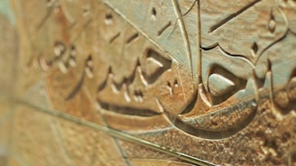 Detailed Arabic calligraphy carved into a textured, stone-like surface with intricate patterns and curves. The image captures the fine details and craftsmanship involved in creating this art form.