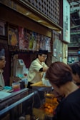 A food vendor in a busy market setting is wearing a traditional-style hat and serving customers. The stall is decorated with colorful posters displaying various food items. There are containers and utensils on the counter, and the vendor is handling cups.