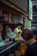 A food vendor in a busy market setting is wearing a traditional-style hat and serving customers. The stall is decorated with colorful posters displaying various food items. There are containers and utensils on the counter, and the vendor is handling cups.