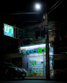 A small illuminated pharmacy storefront is present with signs in Thai script. The interior is well-lit, revealing various products displayed on shelves. A car is parked in front of the store under a streetlamp, and numerous tangled electrical cables run above.