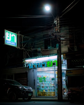 A small illuminated pharmacy storefront is present with signs in Thai script. The interior is well-lit, revealing various products displayed on shelves. A car is parked in front of the store under a streetlamp, and numerous tangled electrical cables run above.