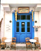 A quaint café with a vibrant blue door flanked by hanging dried gourds. Two small white tables accompanied by wooden chairs are placed in front. Greek signage is displayed above the door and on the wall, with a list of drinks including rakomelo and cocktails.