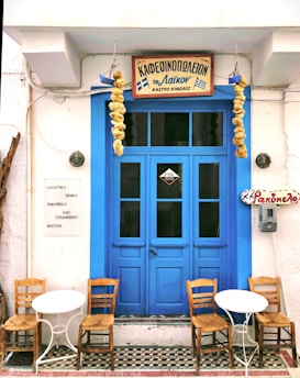 A quaint café with a vibrant blue door flanked by hanging dried gourds. Two small white tables accompanied by wooden chairs are placed in front. Greek signage is displayed above the door and on the wall, with a list of drinks including rakomelo and cocktails.