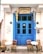 A quaint café with a vibrant blue door flanked by hanging dried gourds. Two small white tables accompanied by wooden chairs are placed in front. Greek signage is displayed above the door and on the wall, with a list of drinks including rakomelo and cocktails.