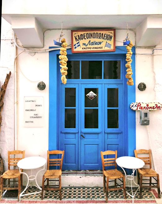 A quaint café with a vibrant blue door flanked by hanging dried gourds. Two small white tables accompanied by wooden chairs are placed in front. Greek signage is displayed above the door and on the wall, with a list of drinks including rakomelo and cocktails.