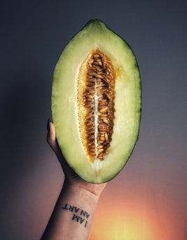 A hand holds a half-cut melon, showcasing its vibrant green rind and the pattern of seeds and fibrous matter in the center. The hand has a tattoo that reads 'I AM AN ART' on the wrist, and the background is a soft gradient transitioning from dark blue to a warm orange hue.
