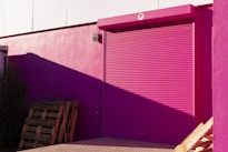 A bright pink roller shutter door is set into a pink wall of a building. The shadow of a structure casts a darker shade on part of the wall. Wooden pallets are stacked against the building, with some vegetation visible to the side. The overall scene is illuminated by sunlight, creating stark contrasts and shadows.