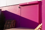 A bright pink roller shutter door is set into a pink wall of a building. The shadow of a structure casts a darker shade on part of the wall. Wooden pallets are stacked against the building, with some vegetation visible to the side. The overall scene is illuminated by sunlight, creating stark contrasts and shadows.