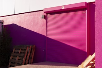 A bright pink roller shutter door is set into a pink wall of a building. The shadow of a structure casts a darker shade on part of the wall. Wooden pallets are stacked against the building, with some vegetation visible to the side. The overall scene is illuminated by sunlight, creating stark contrasts and shadows.
