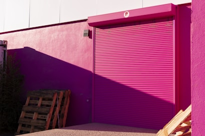 A bright pink roller shutter door is set into a pink wall of a building. The shadow of a structure casts a darker shade on part of the wall. Wooden pallets are stacked against the building, with some vegetation visible to the side. The overall scene is illuminated by sunlight, creating stark contrasts and shadows.