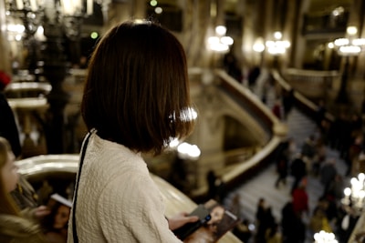 A woman with short, brown hair is standing on a balcony in an ornate, grand interior space with lit chandeliers and a large stairway in the background. She is holding a phone and a brochure. Other people are seen in the background moving up and down the stairs.