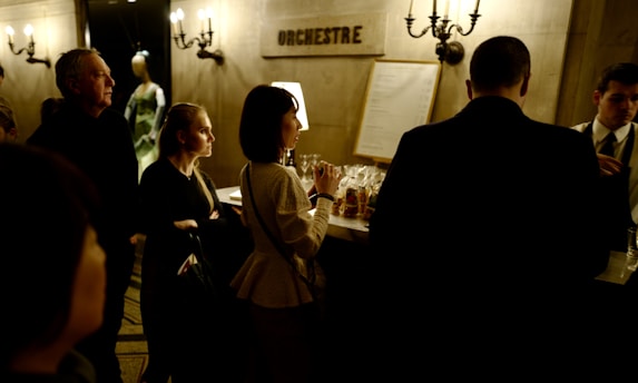 A group of people stands at a dimly lit counter, possibly a bar or ticket counter. A sign reading 'Orchestre' is visible on the wall. The scene conveys a formal or cultural gathering, with individuals dressed in smart attire. The lighting creates a warm and intimate atmosphere.