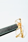 A pair of gold-rimmed glasses is resting on a textured black cylinder against a light background. The glasses have clear lenses and a shiny appearance, while the cylinder has a sparkling surface.