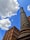 A tall brick chimney stands prominently against a bright blue sky with scattered clouds. Next to it, a multi-story building features a combination of red and tan brickwork with numerous windows.
