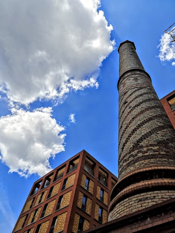 A tall brick chimney stands prominently against a bright blue sky with scattered clouds. Next to it, a multi-story building features a combination of red and tan brickwork with numerous windows.