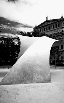 A contemporary, curved metal sculpture stands prominently in the foreground. Behind it, an ornate, classical-style building is visible with detailed architectural elements such as columns and statues. There are trees and parked cars in the background, and the sky appears partly cloudy.