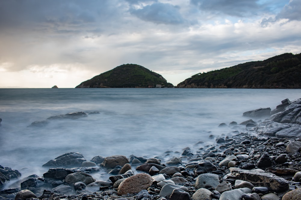 rocky shore with mountain in distance under white clouds and blue sky during daytime