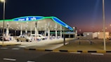 A modern gas station is pictured at dusk with a bright green and blue canopy. Several vehicles, including a white van and a pickup truck, are parked at the pumps. The station is well-lit with numerous overhead lights and features multiple fuel dispensers. The pavement is marked with yellow and black curbs, and the surrounding area appears clean and organized.