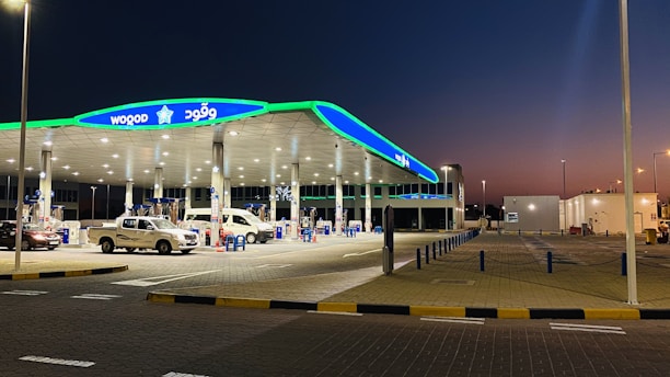 A modern gas station is pictured at dusk with a bright green and blue canopy. Several vehicles, including a white van and a pickup truck, are parked at the pumps. The station is well-lit with numerous overhead lights and features multiple fuel dispensers. The pavement is marked with yellow and black curbs, and the surrounding area appears clean and organized.