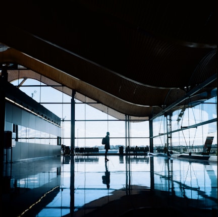 A silhouetted figure is walking through a spacious, modern airport terminal with large glass windows and high ceilings. Natural light reflects off the shiny tiled floor, creating an open and airy atmosphere.