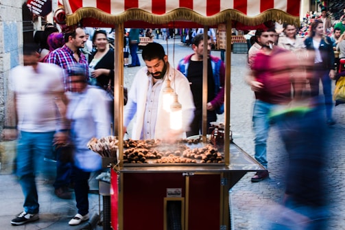 A street vendor stands at a small, brightly lit cart selling roasted chestnuts. The scene is bustling with blurred motion as people walk past quickly, indicating a lively urban setting. The vendor is focused on his work amidst the activity, providing a contrast between his stillness and the surrounding movement.
