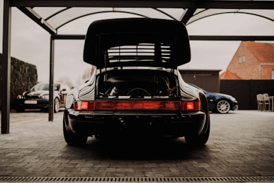 A black Porsche sports car is parked in a covered area with its rear engine compartment open. The surrounding area includes other parked cars, a building with orange rooftops, and some outdoor furniture on a paved surface.