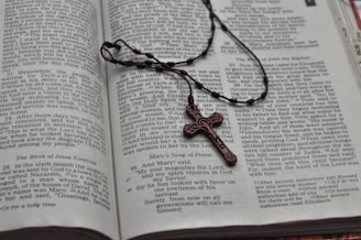 An open book displaying text from the Bible with a wooden rosary cross laid across the pages. The text on the visible pages includes chapters and verses from the Gospel of Luke, specifically 'The Birth of John the Baptist Foretold' and 'Mary's Song of Praise'. The wooden cross is an integral part of the rosary, symbolizing Christian religious devotion.