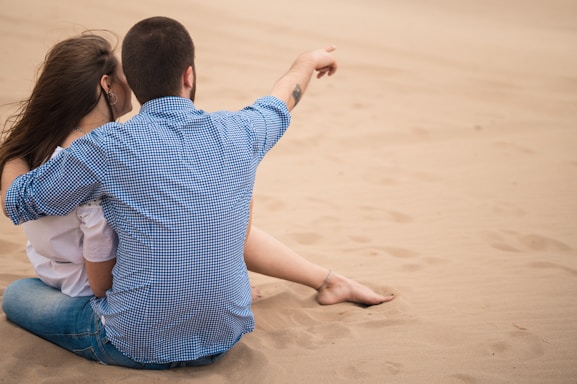A couple sits closely together on a sandy beach. The man has his arm around the woman and is pointing towards something in the distance. Both are wearing casual clothes, with the man in a checkered shirt and the woman in a white top.