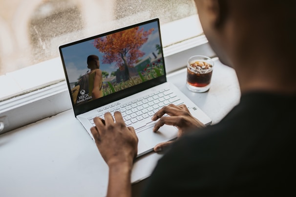 A person is sitting at a table using a white laptop. The laptop screen displays a vibrant video game scene featuring a character walking towards a tree with red leaves. Beside the laptop, there is a glass of dark beverage with ice.