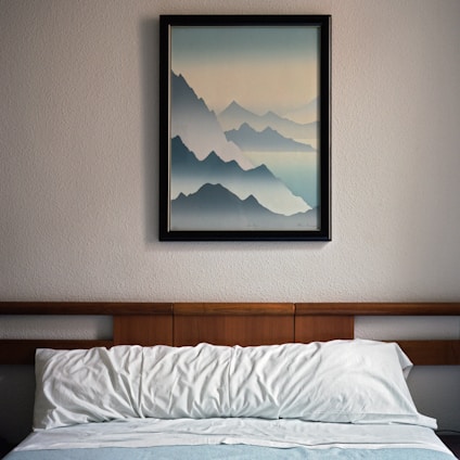 A neatly made bed with white pillows and a blue blanket is situated against a wall. Above the bed, a framed painting depicts layers of blue mountains fading into the horizon, creating a serene and tranquil atmosphere. The bed's headboard is made of wood, complementing the simple and clean decor of the room.