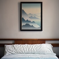 A neatly made bed with white pillows and a blue blanket is situated against a wall. Above the bed, a framed painting depicts layers of blue mountains fading into the horizon, creating a serene and tranquil atmosphere. The bed's headboard is made of wood, complementing the simple and clean decor of the room.