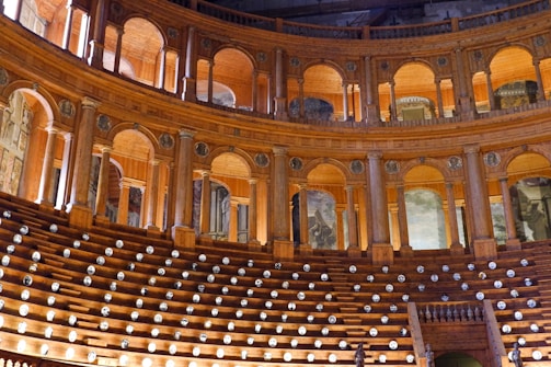 An opulent and historical theater interior featuring wooden arches and seating arranged in a semicircular layout. The architecture is ornate with intricate detailing and paintings adorning the walls. Soft lighting creates a warm ambiance, highlighting the craftsmanship of the wooden structures.