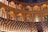 An opulent and historical theater interior featuring wooden arches and seating arranged in a semicircular layout. The architecture is ornate with intricate detailing and paintings adorning the walls. Soft lighting creates a warm ambiance, highlighting the craftsmanship of the wooden structures.