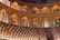 An opulent and historical theater interior featuring wooden arches and seating arranged in a semicircular layout. The architecture is ornate with intricate detailing and paintings adorning the walls. Soft lighting creates a warm ambiance, highlighting the craftsmanship of the wooden structures.