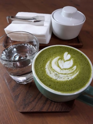 A cup of matcha latte with intricate latte art design on the surface, placed on a wooden coaster. Beside it, there is a glass of water and a white sugar bowl with a lid. In the background, there are white napkins topped with a metal tong on another wooden tray.