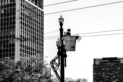 A worker is elevated in a bucket truck, performing maintenance on a streetlight or power line. The scene is urban, with large buildings and trees visible in the background. The image is in black and white, emphasizing the contrast between the structures and the sky.