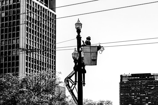 A worker is elevated in a bucket truck, performing maintenance on a streetlight or power line. The scene is urban, with large buildings and trees visible in the background. The image is in black and white, emphasizing the contrast between the structures and the sky.