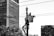 A worker is elevated in a bucket truck, performing maintenance on a streetlight or power line. The scene is urban, with large buildings and trees visible in the background. The image is in black and white, emphasizing the contrast between the structures and the sky.