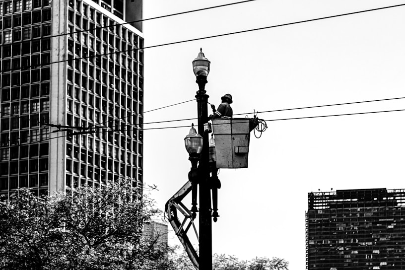 A worker is elevated in a bucket truck, performing maintenance on a streetlight or power line. The scene is urban, with large buildings and trees visible in the background. The image is in black and white, emphasizing the contrast between the structures and the sky.