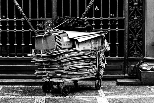 A stack of flattened cardboard boxes and papers are piled on a cart with wheels, positioned in front of a wrought iron gate. The gate features ornate designs, and there is a wheelchair accessibility symbol visible. The scene conveys a sense of urban life and recycling.