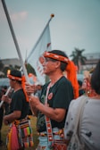 A group of people dressed in traditional attire participate in a cultural or ceremonial event outdoors. One individual in the foreground holds a flag, wearing a headband and attire featuring vibrant colors and patterns. Other participants in similar clothing are visible in the background.
