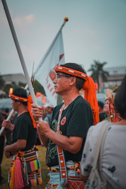 A group of people dressed in traditional attire participate in a cultural or ceremonial event outdoors. One individual in the foreground holds a flag, wearing a headband and attire featuring vibrant colors and patterns. Other participants in similar clothing are visible in the background.