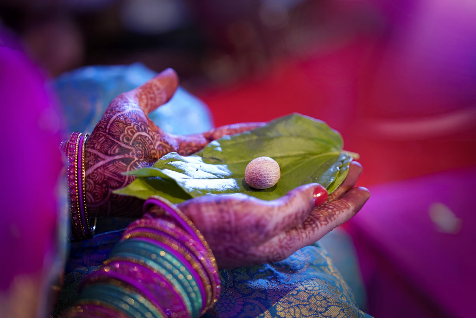 Hennaed hands in stacked bangles holding a betel leaf with an areca nut resting on it.
