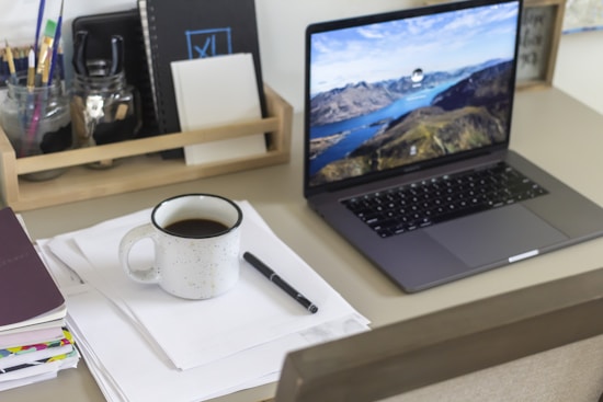 A workspace featuring a wooden organizer holding notebooks and jars of stationery items, beside a stack of papers with a pen and a coffee mug. A laptop displaying a scenic landscape is open on the desk.